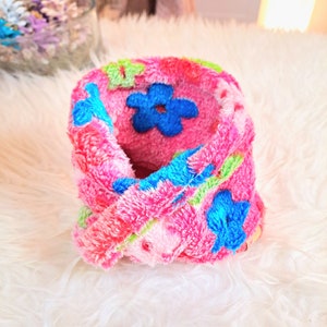 May include: Pink fuzzy neck warmer with a floral pattern in blue, green, and pink. The neck warmer is folded in half and has a flower design on the front.