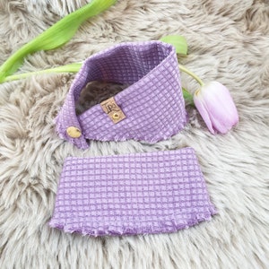 Può includere: Un cerchietto in tessuto a nido d'ape viola con un bordo sfrangiato e un piccolo cartellino marrone con la scritta "Lro" su di esso.