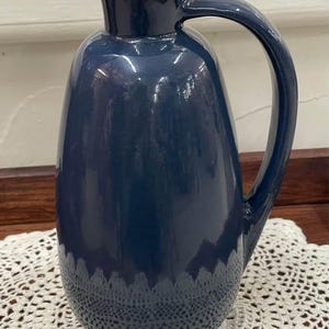 May include: A glossy, deep navy blue ceramic pitcher with a smooth, rounded body, a curved handle on the side, and a small pouring spout at the top. The pitcher is topped with a light brown, cylindrical cork stopper that fits snugly into the opening.