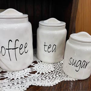 May include: Three white ceramic canisters with lids, labeled "coffee", "tea", and "sugar" in black script. The canisters are arranged on a white lace doily, suggesting a kitchen or dining setting.