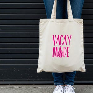 Puede incluir: Una bolsa de tela blanca con el texto "VACAY MODE" en letras rosas y un gr&aacute;fico de pi&ntilde;a rosa.