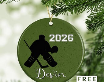 Peut inclure: Ornement de Noël rond vert olive avec une silhouette noire de gardien de but de hockey. L'année "2026" est imprimée en blanc, avec le nom "Devin" en cursive. Une ficelle dorée est attachée pour l'accrocher. Le texte "FREE GIFT BOX" est en bas à droite.