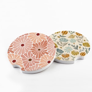May include: Two car coasters with floral designs. One coaster features a pattern of pink and peach flowers with brown centers. The other has a cream background with blue, yellow, and brown floral and leaf patterns.