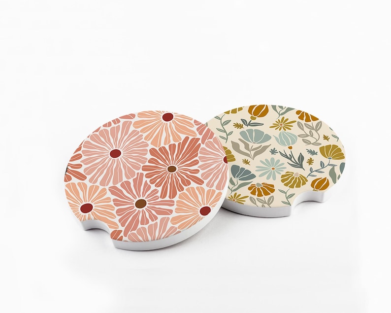 May include: Two car coasters with floral designs. One coaster features a pattern of pink and peach flowers with brown centers. The other has a cream background with blue, yellow, and brown floral and leaf patterns.