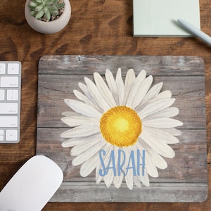 May include: A white daisy with a yellow center on a woodgrain background. The name "SARAH" is written in blue below the flower.