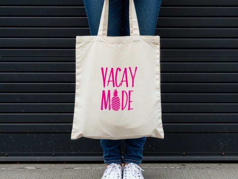 Puede incluir: Una bolsa de tela blanca con el texto "VACAY MODE" en letras rosas y un gr&aacute;fico de pi&ntilde;a rosa.