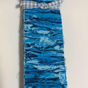 May include: A stack of blue fabric strips with a marbled pattern, tied with a light blue and white gingham ribbon. The fabric has a mix of light and dark blue hues with black accents, creating a textured appearance. The edges of the fabric are frayed.