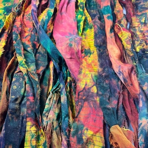 May include: Close-up of colorful fabric strips in shades of yellow, orange, pink, green, and blue. The fabric appears to be hand-dyed or tie-dyed, with a textured, slightly frayed appearance. The strips are layered and overlapping, creating a vibrant, abstract design.