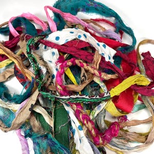 May include: A collection of assorted fabric strips in various colors including teal, magenta, yellow, and white with blue polka dots. The strips feature frayed edges, varying textures, and some braided sections, creating a tangled, layered pile.