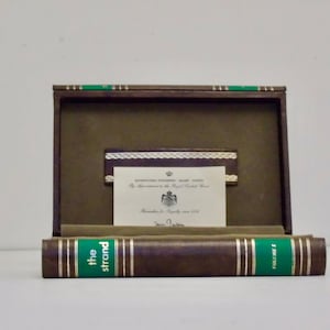 Puede incluir: Un libro encuadernado en cuero marrón titulado "The Strand" con letras doradas y detalles verdes. El libro está en una caja marrón con un forro verde y una tarjeta en el interior que dice "The Strand Magazine is the Royal Standard Book".