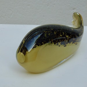 May include: A decorative glass sculpture in the shape of a whale. The sculpture features a combination of colors, including a light yellow base and a dark brown speckled pattern on the top. The whale has a curved tail and a smooth, glossy finish.