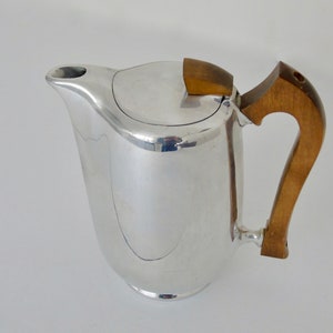 Puede incluir: Una cafetera plateada con asa y tapa de madera. El asa es curvada y está unida al lateral de la cafetera. La tapa también es plateada y tiene un adorno de madera. La cafetera tiene un pico para verter.