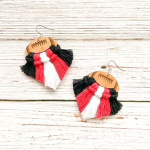 May include: A pair of wooden football earrings with red, white, and black tassels. The earrings are made of wood and have a football design. The tassels are made of yarn and are tied to the earrings.