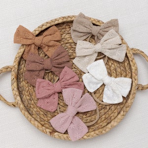 May include: A set of six hair bows in various shades of brown, pink, and white. The bows are made of a delicate, embroidered fabric with a floral pattern. The bows are arranged in a circular pattern on a woven basket.