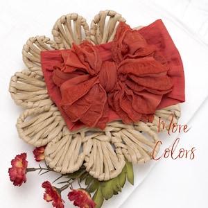 May include: A rust-colored fabric headband featuring a large, ruffled bow detail. The headband is placed on a woven tan flower-shaped base with small red flowers at the bottom. The text "More Colors" is printed in a script font on the right side.