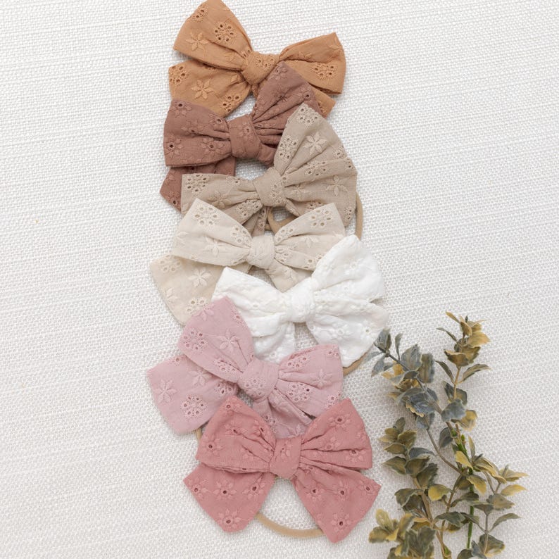 May include: Set of six hair bows in various shades of brown, pink, and white. Each bow is made of a delicate, eyelet fabric with a floral pattern. The bows are tied with a simple knot and are attached to a nude elastic headband.