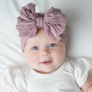 May include: A dusty rose-colored headwrap featuring a large, textured, multi-layered fabric bow on the front. The accessory is made of a soft, stretchy material that fits snugly around the head, showcasing a gathered design and a ruffled edge.