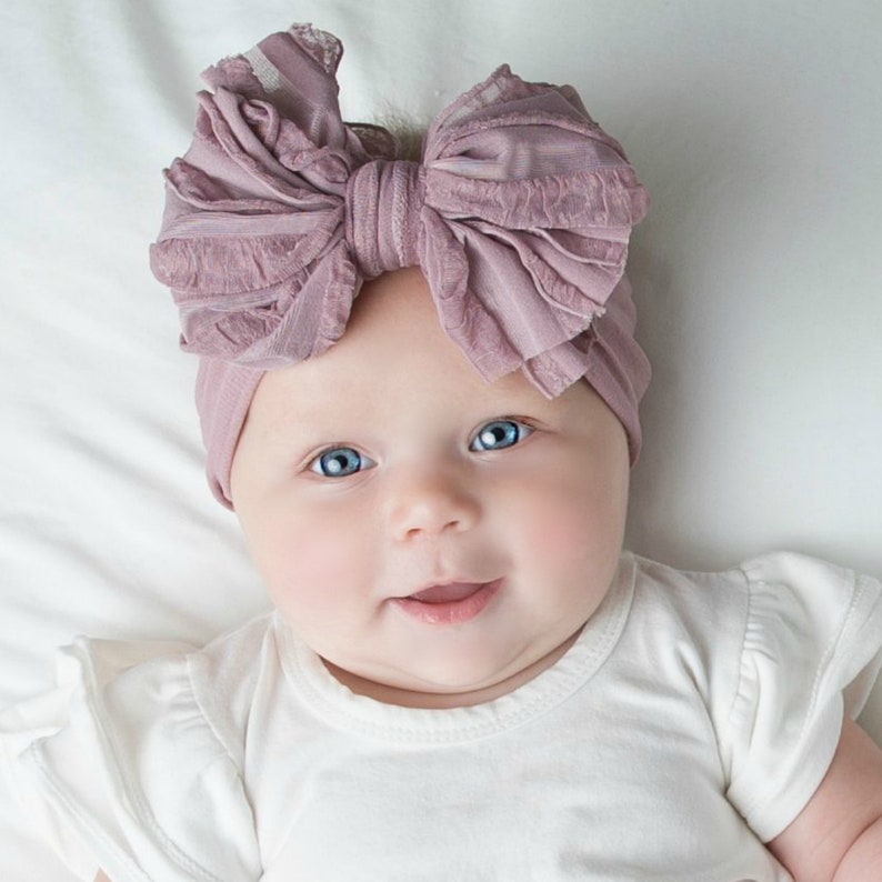 May include: A dusty rose-colored headwrap featuring a large, textured, multi-layered fabric bow on the front. The accessory is made of a soft, stretchy material that fits snugly around the head, showcasing a gathered design and a ruffled edge.
