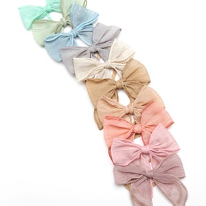 May include: A set of ten hair bows in various pastel colors, made from sheer fabric with a subtle shimmer. The bows are arranged in a line, with the lightest color at the top and the darkest color at the bottom. Each bow is attached to a nude elastic headband.
