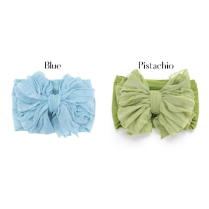May include: Two fabric headbands featuring large, ruffled bows. One headband is light blue, labeled "Blue," and the other is a muted green, labeled "Pistachio." Both items have a textured, layered appearance with a central knot detail on the bow.