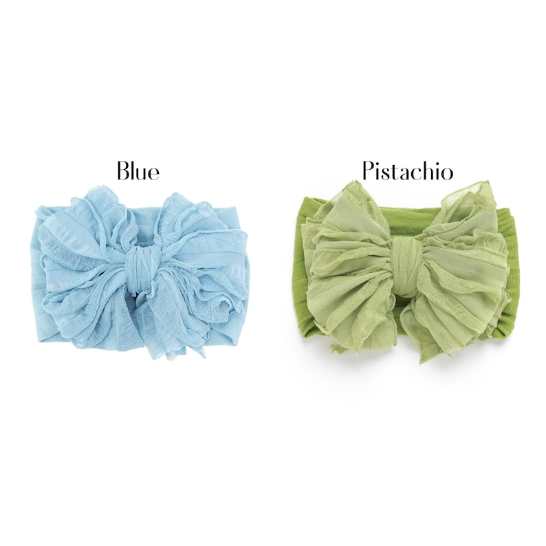 May include: Two fabric headbands featuring large, ruffled bows. One headband is light blue, labeled "Blue," and the other is a muted green, labeled "Pistachio." Both items have a textured, layered appearance with a central knot detail on the bow.