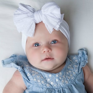 May include: A white fabric headband featuring a large, textured, ruffled bow on top. The headband is worn with a light blue denim-style top that has short ruffled sleeves and a delicate white floral embroidery pattern across the chest area.