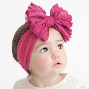 May include: A dark pink fabric headband featuring a large, multi-layered bow made of ruffled, semi-sheer material. The headband is a solid, stretchy knit fabric that sits securely around the head, with the prominent bow positioned on the top side.