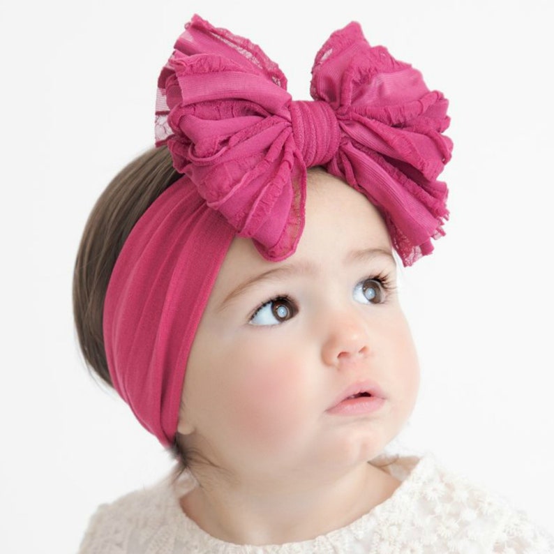 May include: A dark pink fabric headband featuring a large, multi-layered bow made of ruffled, semi-sheer material. The headband is a solid, stretchy knit fabric that sits securely around the head, with the prominent bow positioned on the top side.
