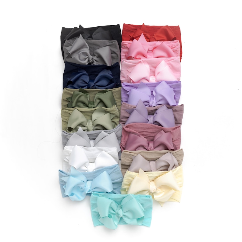Peut inclure: Une collection de bandeaux pour b&eacute;b&eacute;s de diff&eacute;rentes couleurs, dont le noir, le gris, le marine, le vert olive, le rose, le lavande et le bleu clair. Chaque bandeau est orn&eacute; d'un grand n&oelig;ud d&eacute;coratif. Les bandeaux sont dispos&eacute;s verticalement sur fond blanc.