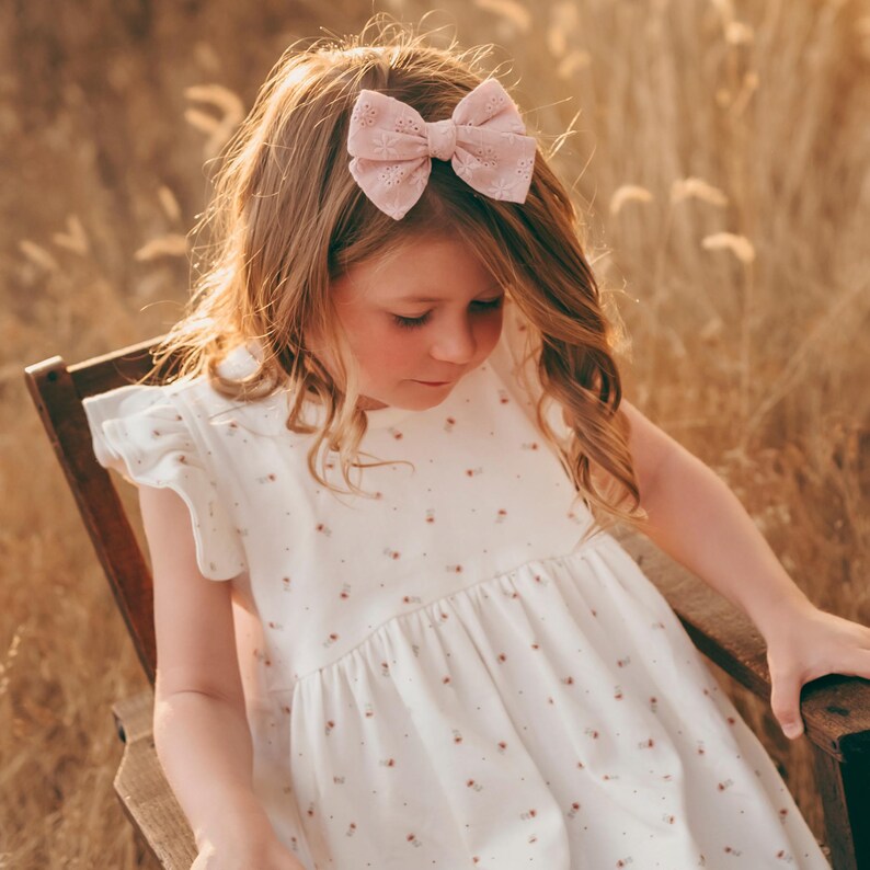 May include: A young girl wearing a white dress with a floral pattern and a pink bow in her hair. She is sitting on a wooden chair in a field of tall grass.