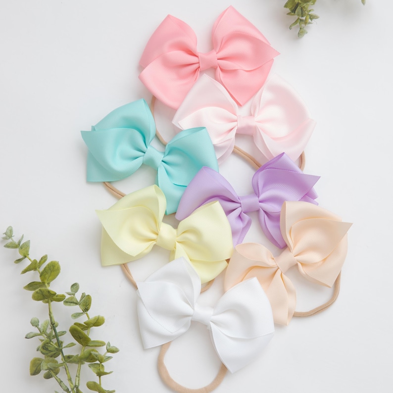 May include: A set of 8 pastel colored hair bows on nude headbands. The bows are made of satin ribbon and are arranged in a circle. The colors of the bows are pink, light blue, lavender, yellow, peach, and white.