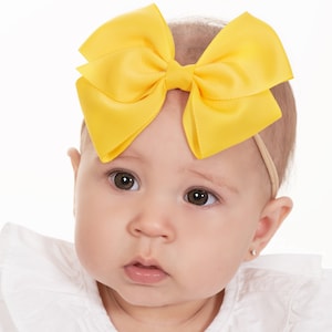 May include: A baby wearing a white shirt with a large yellow bow headband.