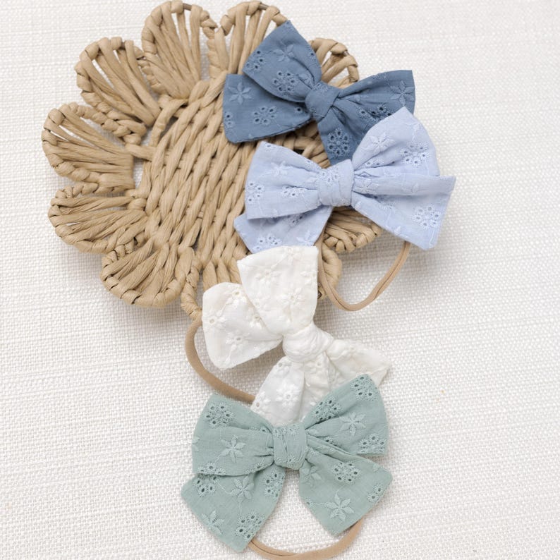 May include: Four handmade hair bows with elastic bands. The bows are made of white, light blue, and blue fabric with a delicate floral pattern.
