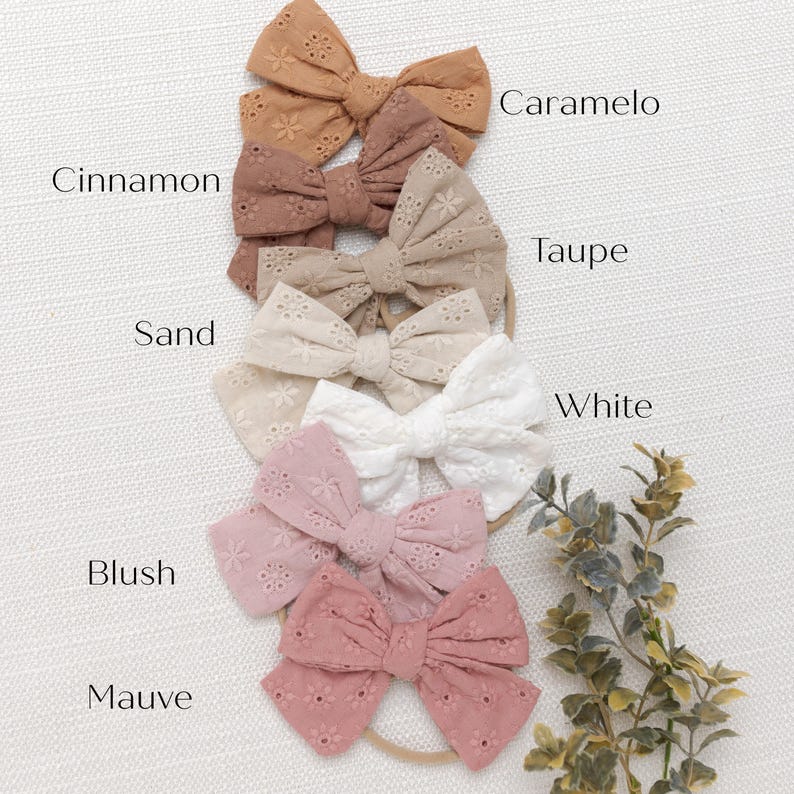 May include: A set of 10 hair bows in various shades of brown, pink, and white. The bows are made of a delicate, eyelet fabric and are arranged in a vertical line. The bows are labeled with the following names: Caramel, Taupe, White, Blush, Mauve, Cinnamon, Sand.