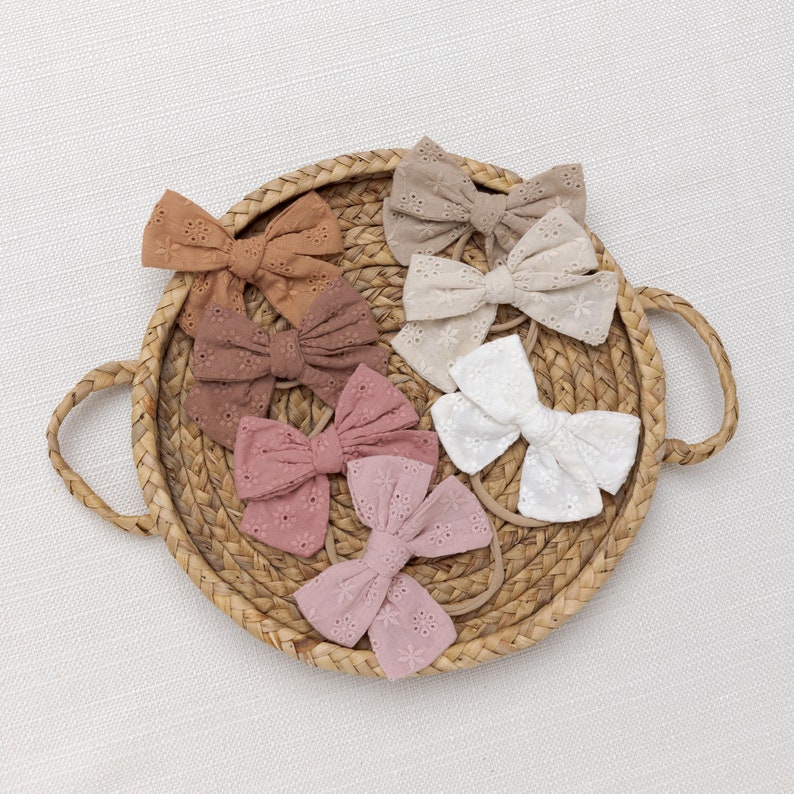 May include: A set of six hair bows in various shades of brown, pink, and white. The bows are made of a delicate, embroidered fabric with a floral pattern. The bows are arranged in a circular pattern on a woven basket.