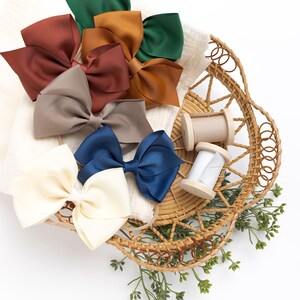 May include: A wicker basket filled with a variety of fabric bows in different colors, including brown, beige, green, blue, and white. There are also two spools of thread in the basket.