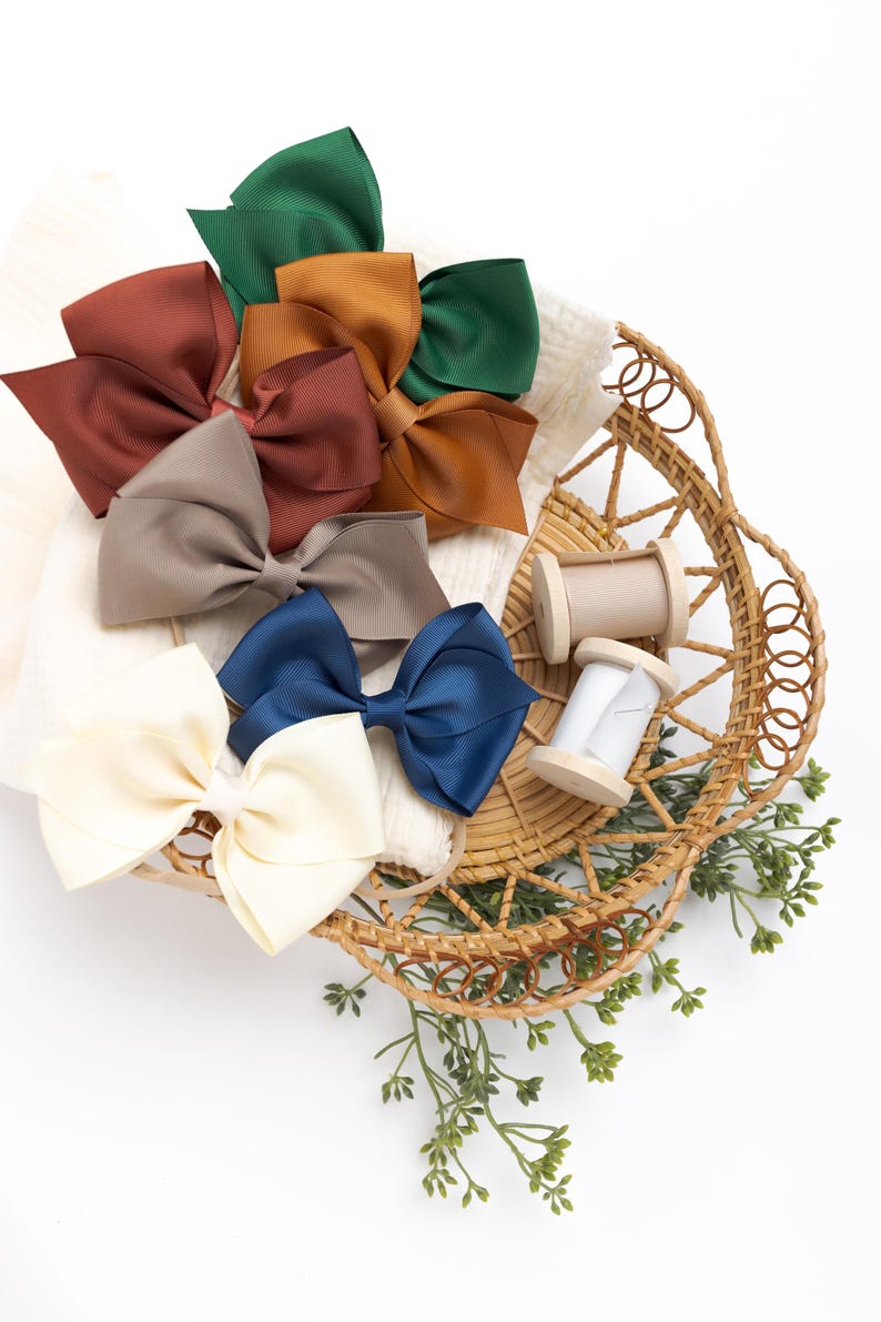 May include: A wicker basket filled with a variety of fabric bows in different colors, including brown, beige, green, blue, and white. There are also two spools of thread in the basket.