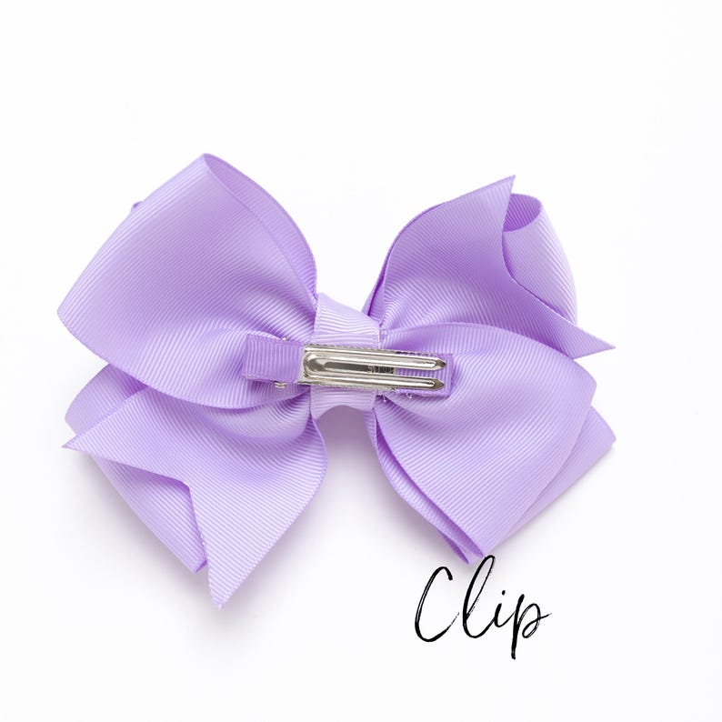 Peut inclure: Un n&oelig;ud de cheveux lilas avec une pince en m&eacute;tal argent&eacute;. Le n&oelig;ud est fait d'un large ruban gros-grain et est nou&eacute; en forme de n&oelig;ud classique. Le mot "Clip" est &eacute;crit en cursive en bas &agrave; droite.