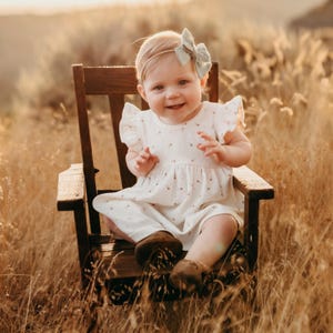 May include: A young child wearing a white dress with a floral pattern and a light blue bow in their hair sits on a wooden chair. The child is smiling and looking at the camera. The chair is in a field of tall grass.