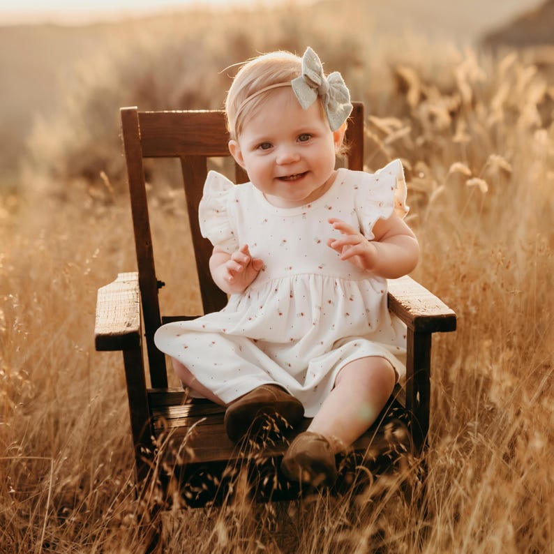 May include: A young child wearing a white dress with a floral pattern and a light blue bow in their hair sits on a wooden chair. The child is smiling and looking at the camera. The chair is in a field of tall grass.
