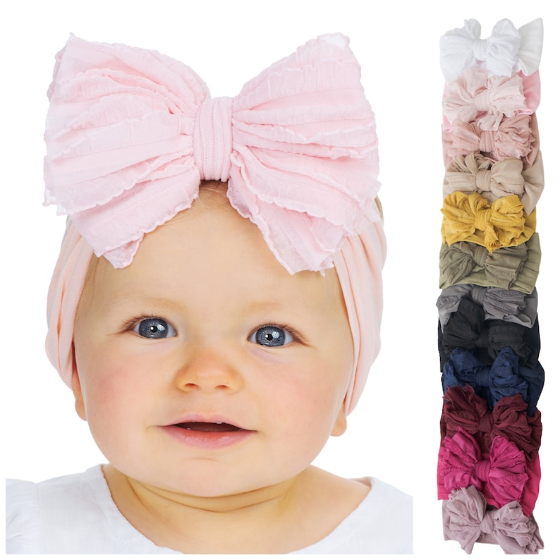 May include: A light pink fabric headband featuring a large, ruffled bow on top. Beside the headband is a vertical stack of eleven additional headbands in various solid colors, including white, beige, mustard yellow, olive green, black, and navy.