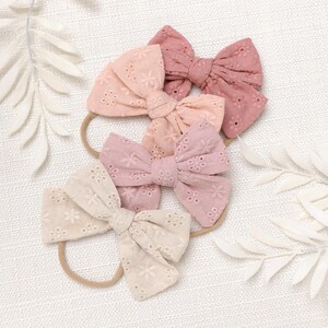 May include: Three pink and beige fabric hair bows with eyelet detailing. The bows are tied with a knot in the center and are attached to elastic headbands.