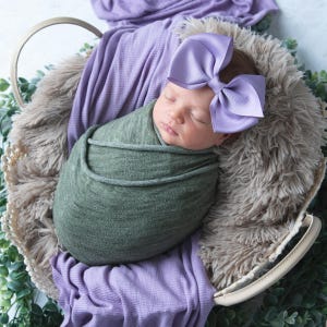 May include: A newborn baby is sleeping in a basket, wrapped in a green swaddle and wearing a purple bow.