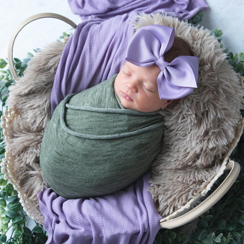 May include: A newborn baby is sleeping in a basket, wrapped in a green swaddle and wearing a purple bow.