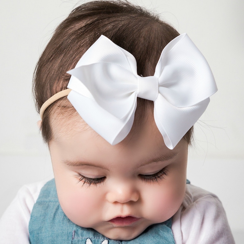 May include: A baby wearing a white headband with a large white bow. The headband is made of elastic and the bow is made of satin.