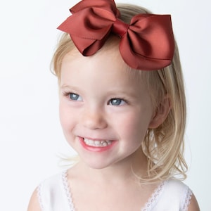 May include: A young girl with blonde hair is wearing a rust-colored bow headband. She is smiling and looking to the side.