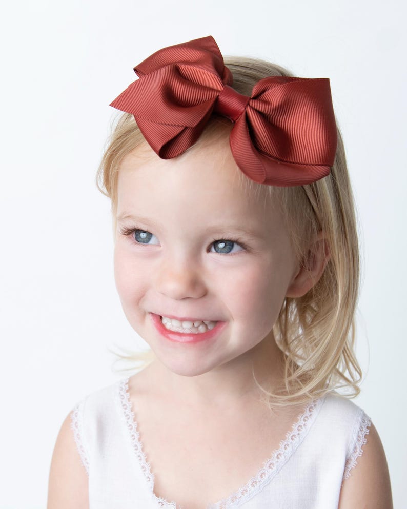 May include: A young girl with blonde hair is wearing a rust-colored bow headband. She is smiling and looking to the side.