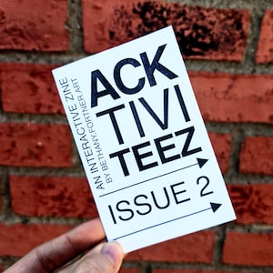 May include: A white rectangular card with the text "ACK TIVITEEZ ISSUE 2" in black, held against a red brick wall. The card also includes the text "AN INTERACTIVE ZINE BY BETHANY.FORTNER.ART".