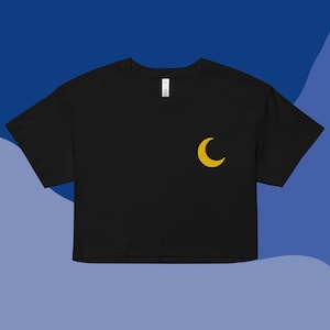 Pode incluir: Top preto curto com uma lua crescente amarela bordada na frente. A camisa tem mangas curtas e gola redonda. O fundo é um gradiente de tons de azul.
