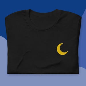 Pode incluir: Camiseta preta com uma lua crescente amarela bordada no peito esquerdo. A camisa é feita de um material macio e está cuidadosamente dobrada. O fundo é um gradiente de azul.